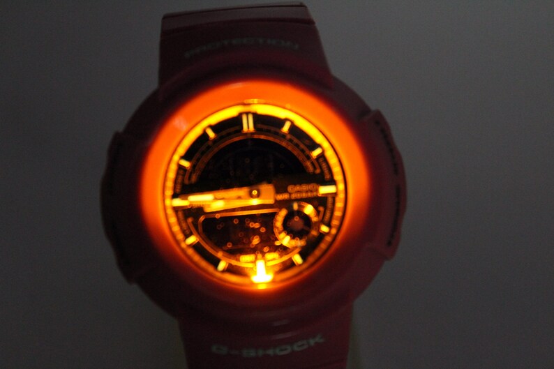 Puede incluir: Un reloj G-Shock rosa con una esfera negra y una luz de fondo naranja brillante. El reloj tiene una pantalla digital y una correa negra.