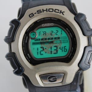 May include: A silver and black G-Shock digital watch with a green display. The watch face shows the date, time, and day of the week. The watch has a black strap.
