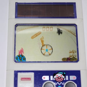 May include: A white and blue handheld electronic game with a clown face on the front. The game is called "Funky Pierrot" and is made by Casio. The screen shows a game over message and a clown juggling rings.