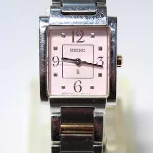 Puede incluir: Un reloj Seiko de tono plateado y dorado con una esfera cuadrada y una esfera rosa. El reloj tiene una correa plateada y un fondo blanco con números y manecillas negros. La esfera del reloj también tiene el texto "SEIKO" y "JAPAN  (M01)" impreso en él.