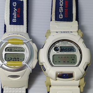 Pode incluir: Dois relógios digitais com caixas de resina branca e bege e pulseiras de tecido azul. O relógio da esquerda é um Baby-G com aro prateado e detalhes amarelos. O da direita é um G-Shock com um visor exibindo o texto PASSPC.