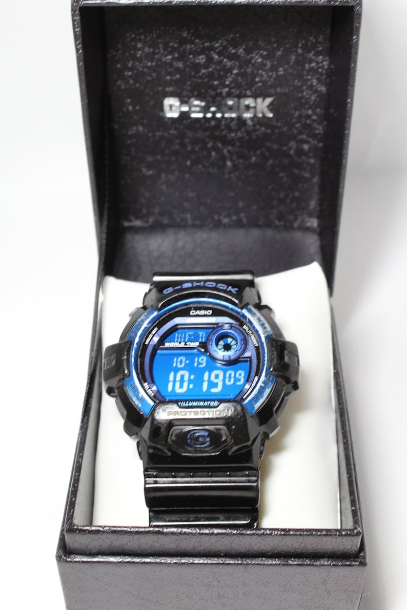 May include: Black G-Shock digital watch with a blue and black bezel, displayed in a black box. The watch face shows the date and time. The watch band is black.