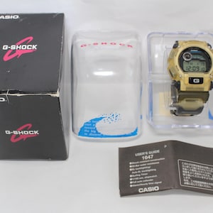 Puede incluir: Un reloj Casio G-Shock dorado con una banda negra y una pantalla digital. El reloj está en su embalaje original, que incluye una guía del usuario. La esfera del reloj muestra la hora, la fecha y el día de la semana.