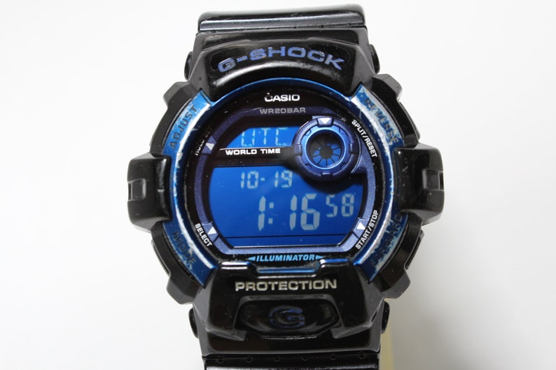 May include: Black and blue G-Shock digital watch with a blue digital display showing the time. The watch has multiple buttons and text including "G-SHOCK", "CASIO", and "WR20BAR". The watch face displays "10-19" and "1:16:58".