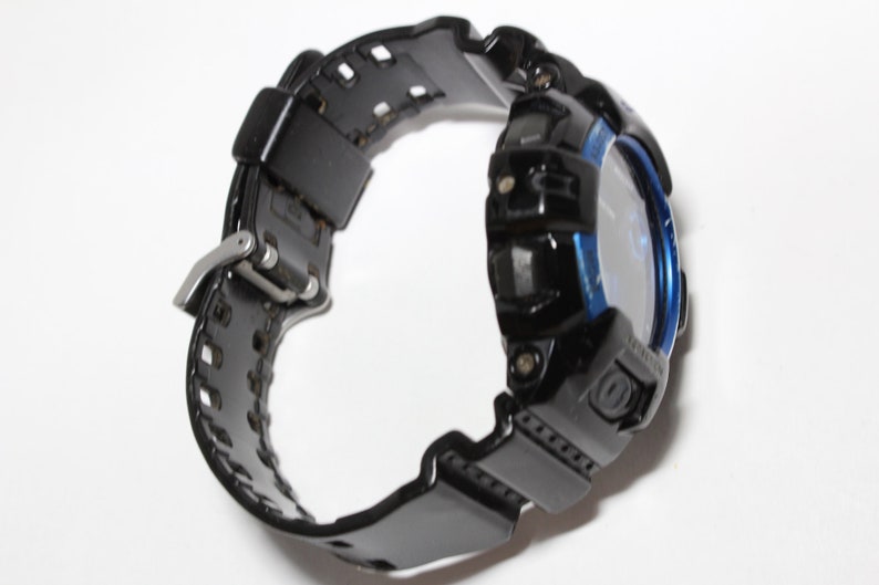 May include: Black digital watch with a blue bezel and a black strap. The watch has a silver buckle and a textured strap design. The watch is designed for sports and outdoor activities.