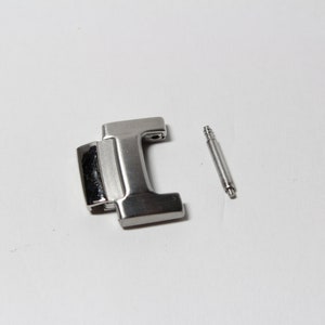 Op de afbeelding: Een zilveren horlogebandgesp met een veerpen. De gesp is rechthoekig met een glad oppervlak en een klein gaatje aan de zijkant. De veerpen is een dunne metalen staaf met een afgerond uiteinde.