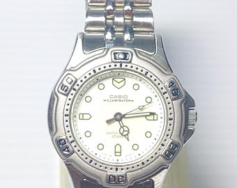May include: A silver Casio Illuminator watch with a white face and a stainless steel strap. The watch has a round face with luminous markers and hands, and the text "WATER 20 BAR" is visible. The strap is made of interlocking links.