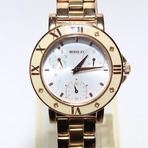 May include: A gold-tone watch with a white face and Roman numeral hour markers. The watch has a day and date display, and a small second hand. The watch is on a gold-tone metal band.