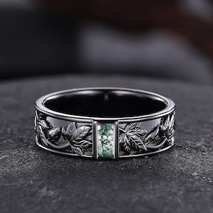 May include: A silver ring with a rectangular green gemstone set in the center. The band features a detailed leaf design. The ring is displayed on a dark, textured surface, highlighting its intricate details and craftsmanship.