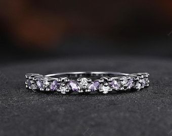 May include: A black band ring with a row of small, clear and purple gemstones. The ring is set on a dark gray surface.