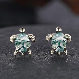 May include: A pair of silver-toned turtle stud earrings. Each earring features a green and white stone set within a turtle-shaped frame. The earrings are displayed on a dark gray surface.