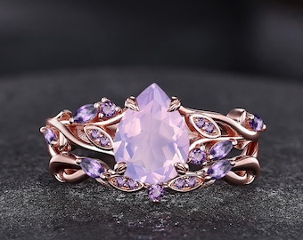 May include: A rose gold ring featuring a central pear-shaped light purple gemstone. The band is designed with intricate metal vines and leaves, accented with small, round, deep purple stones embedded along the branches surrounding the center stone.