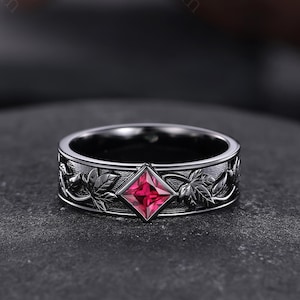 May include: A dark gunmetal grey band ring featuring a central square-cut vibrant red gemstone. The band is decorated with an embossed relief pattern of detailed leaves and vines wrapping around the entire circumference of the metal surface.