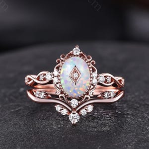 May include: A rose gold ring set featuring an oval-shaped opal center stone with a small diamond-shaped accent in the middle. The setting includes intricate filigree metalwork and small round white stones along the band and the curved wedding band.