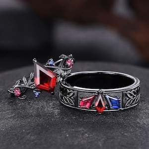 May include: Two black rings with gemstone accents. One ring features a large, red, diamond-shaped stone and smaller pink and blue stones. The other ring has a band with a floral design and red, pink, and blue gemstones.
