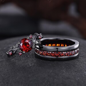 Könnte beinhalten: Zweiteiliges Ring-Set. Der schwarze Bandring hat eine Reihe roter Edelsteine. Der Verlobungsring hat einen großen, ovalen roten Edelstein und ein florales Design. Die Ringe werden auf einer dunklen Oberfläche präsentiert.