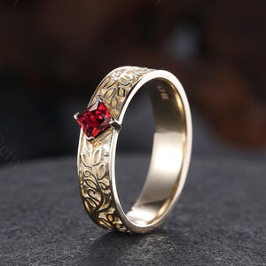 Pode incluir: Um anel de ouro com uma pedra vermelha quadrada. A banda apresenta um intrincado padrão floral. O anel está sobre um fundo escuro, destacando os seus detalhes. O anel é uma joia.