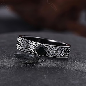 Puede incluir: Un anillo negro con un diseño grabado intrincado y una gema negra cuadrada. El anillo se encuentra sobre una superficie oscura, mostrando sus detalles. La banda presenta un patrón detallado.