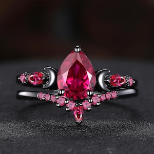 May include: A black ring set featuring a pear-shaped, vibrant red gemstone as the centerpiece. The ring is accented with smaller pink gemstones and crescent moon details, creating a celestial design. The band is black.