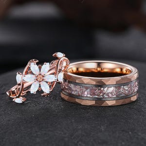 Op de afbeelding: Twee roségouden ringen. Eén ring heeft een bloemenontwerp met witte opaalstenen. De andere ring heeft een gehamerde textuur met een band van roze stenen en een roségouden afwerking. Sieraden.