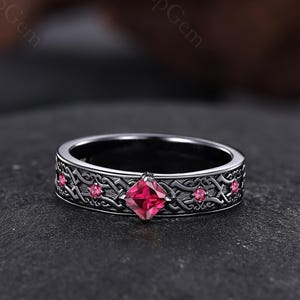 Puede incluir: Un anillo plateado con una gema roja cuadrada en el centro y gemas rojas más pequeñas en los lados. El anillo tiene un intrincado diseño de inspiración celta. El anillo se muestra sobre una superficie oscura y texturizada.