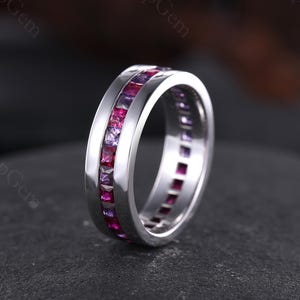 May include: A silver ring with a channel setting, featuring alternating square-cut pink and purple gemstones. The ring is set against a dark background, accentuating the vibrant colours of the gemstones.