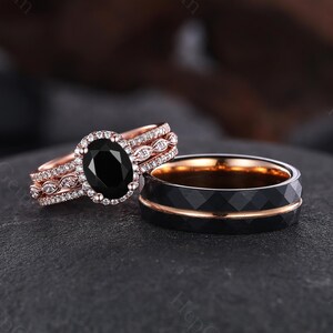 May include: A rose gold engagement ring featuring a large oval black gemstone, encircled by a halo of smaller white stones. A matching black and rose gold wedding band is next to it. The wedding band has a geometric design.