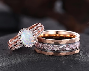 Puede incluir: Dos anillos de oro rosa. Uno presenta una piedra central ovalada de ópalo rodeada por un halo de pequeños diamantes. El segundo tiene una textura martillada con un patrón floral y una banda de piedras rosas. Los anillos se exhiben sobre una superficie oscura.