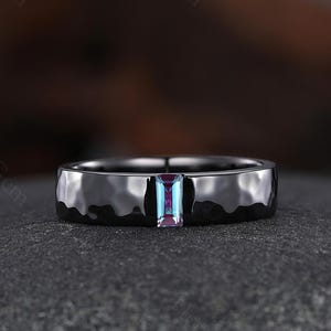 May include: A black hammered-finish ring with a rectangular, multi-colored gemstone. The gemstone displays shades of blue, purple, and pink. The ring is set against a dark, textured background.