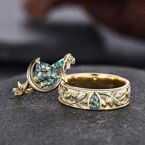 May include: A gold ring set featuring a crescent moon and leaf motif. The engagement ring has a pear-shaped moss agate and small green stones. The matching band displays an engraved leaf pattern with a single pear-shaped moss agate inlay.