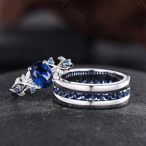 May include: A silver-toned ring set consisting of an engagement ring with an oval-cut blue center stone and leaf-shaped accents, and a matching band featuring a channel-set row of square-cut blue stones encircling the entire circumference.
