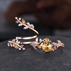 May include: A rose gold ring set featuring a pear-shaped yellow citrine center stone and smaller leaf-shaped accents. The bands are designed with a vine-like motif, embellished with tiny clear and yellow stones set along the metal branches.