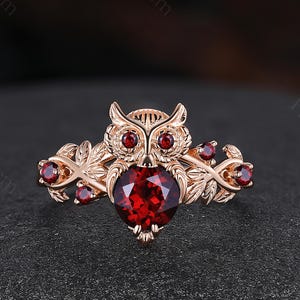 May include: A rose gold ring featuring an owl design with a red garnet. The owl is surrounded by leaves and smaller red garnets.