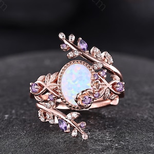 May include: A rose gold ring featuring a central oval opal cabochon surrounded by a beaded border. The band is designed with intricate metal leaf motifs, small clear stones, and tiny purple accent stones set along the branching metal vines.