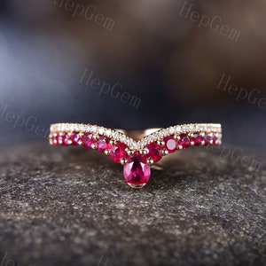 Puede incluir: Un anillo de banda de oro con un diseño en forma de V que presenta una fila de pequeñas piedras de rubí rojo y una piedra de rubí más grande en la parte inferior. El anillo está engastado con diamantes más pequeños a lo largo de la banda.