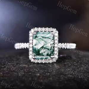 May include: A silver ring with a large, rectangular, green moss agate gemstone surrounded by a halo of smaller diamonds. The ring has a delicate band with smaller diamonds.