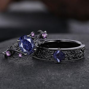 May include: A matching set of rings with a dark metal finish. One ring features a large, round, blue gemstone with a sparkling effect, surrounded by small pink stones and leaf accents. The other ring has a square blue gemstone and leaf pattern.