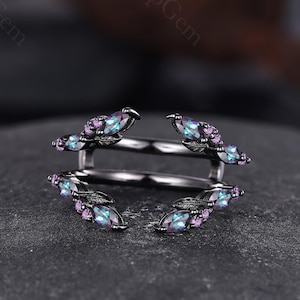 以下が含まれることがあります： 黒い金属製のリングガードで、葉の形をしたアクセントで繋がれた2つのオープンバンドが特徴です。葉には虹色のマーキスカットの石と小さな丸い紫色の石がセットされており、中央のリングを囲むようにデザインされた装飾的なフレームを形成しています。