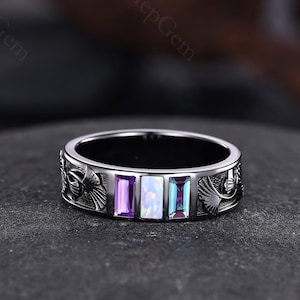 May include: A black metal ring featuring three rectangular stones set in the center: a purple amethyst, a white iridescent opal, and a blue-toned stone. The band is decorated with engraved ginkgo leaf patterns on both sides of the stones.