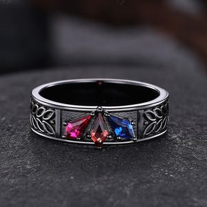 May include: A silver ring with three colorful gemstones: red, pink, and blue. The ring band features a decorative leaf pattern. The gemstones are set in a triangular arrangement, adding a unique touch to the jewelry.