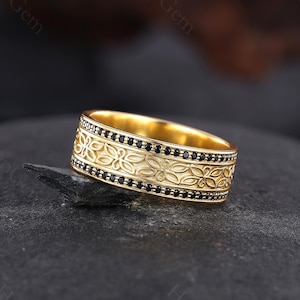 May include: A gold-colored ring with an intricate floral pattern and a row of small black stones. The ring is set on a dark, textured surface, showcasing its detailed design and craftsmanship. The ring's band is approximately 0.5 inches wide.