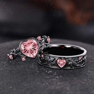 May include: A black metal ring set featuring a prominent heart-shaped pink gemstone on one band and a matching band with a smaller heart-shaped pink gemstone, both adorned with intricate leaf engravings and small pink accent stones throughout.