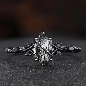 May include: Black engagement ring with a clear, oval-shaped gemstone with black inclusions. The ring band is black and features a twisted design with small black gemstones.