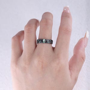 May include: A black ring with a rectangular emerald-colored stone in the center and smaller emerald-colored stones on the sides. The ring has a decorative pattern and is worn on a finger.