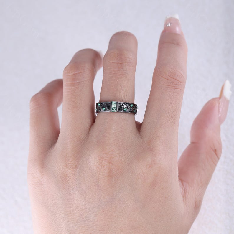 May include: A black ring with a rectangular emerald-colored stone in the center and smaller emerald-colored stones on the sides. The ring has a decorative pattern and is worn on a finger.