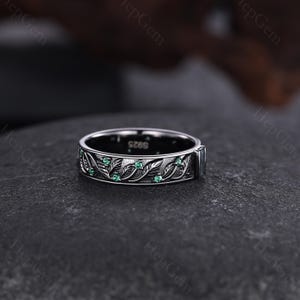 May include: A silver ring with a leaf and vine design, accented with small green gemstones. The ring has a dark background and is likely made of sterling silver. The ring's band features an engraved pattern of leaves and vines.
