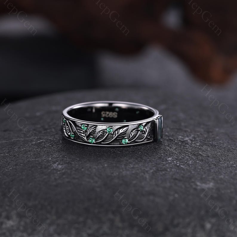 May include: A silver ring with a leaf and vine design, accented with small green gemstones. The ring has a dark background and is likely made of sterling silver. The ring's band features an engraved pattern of leaves and vines.