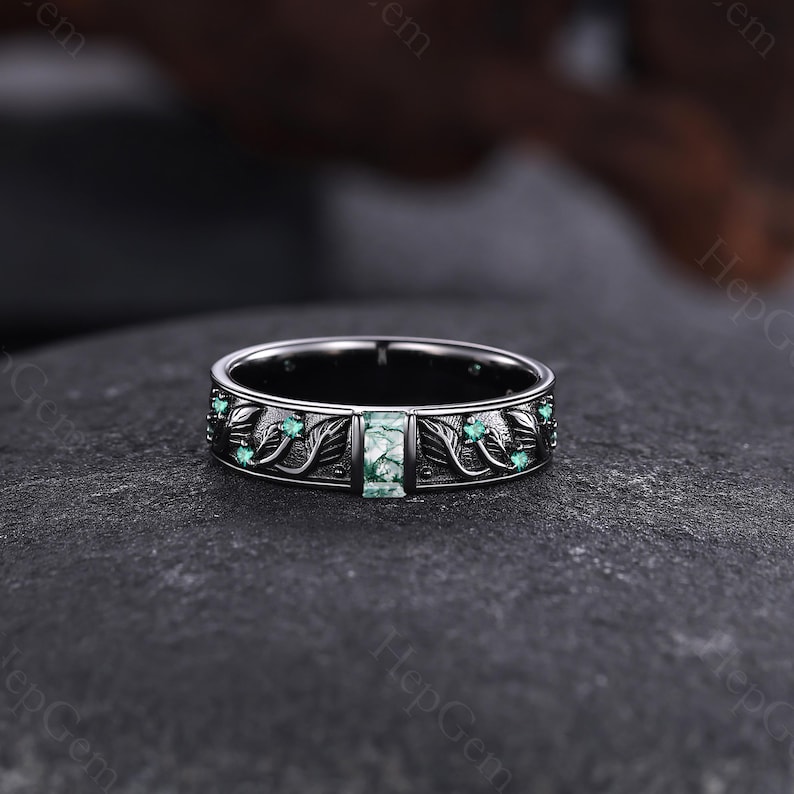 May include: A silver ring with a rectangular green stone at the center. The ring features a detailed leaf and vine design with small green stones. The ring is set against a dark background, highlighting its intricate details.