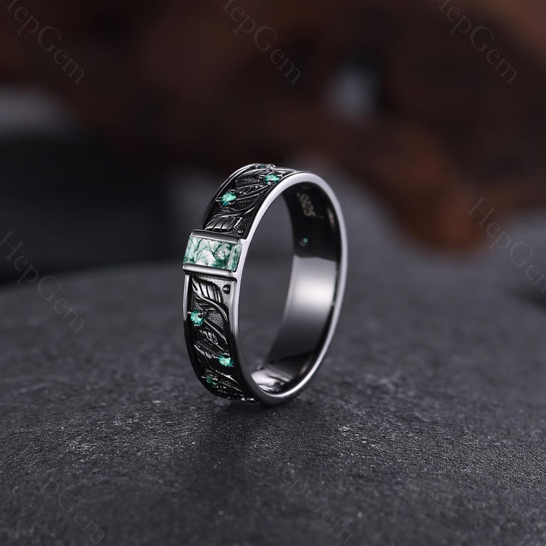 May include: A silver ring with a rectangular green stone and leaf design. The ring has a dark band with engraved leaves and small green stones. The ring is on a dark gray surface.