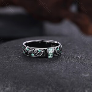 May include: A silver ring with a rectangular green stone at the center. The ring features a leaf and vine design with small green stones. The ring is set on a dark surface.